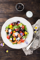 Wall Mural - Fresh salad with smoked salmon, black olives, cherry tomatoes and edible flowers on wooden background. Top view.