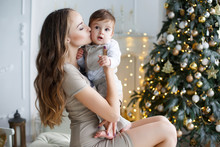 Happy Brunette Woman With Long Hair,holds A Christmas Party, Sitting On A White Sofa Near The Christmas Tree With Their Little Son In Her Arms,Christmas Portrait Of A Mother And Child