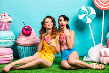 two young cool lady on turquoise background, sitting on the grass and eating popcorn, laughing, best