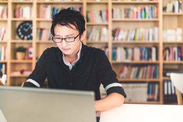 Young pensive Asian man working on laptop at home office or library with serious face, bookshelf blur background with copy space, business or technology concept