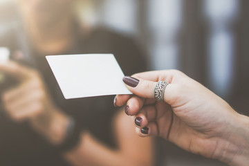 Closeup of instant photo in woman's fingers. Blank white business card in female's hand. Ring on finger. In background is blurred woman holding an instant camera. Film effect,blurred background.