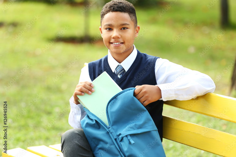 Schoolboy putting book into backpack on bench in park