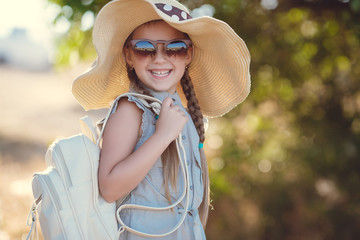 Young traveler girl in a straw hat with a large brim,dark sunglasses,light-blue summer suit,behind carries a blue backpack,posing outdoors in the summer in the countryside amid fields and green bushes