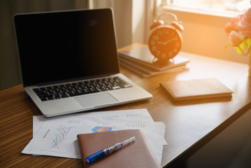 A wooden desk with a laptop, notebook,clock,pen and annual reports in morning,working space.