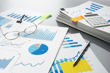 Preparing report. Blue graphs, glasses, calculator and pen.　Business reports and pile of documents on gray reflection background.
