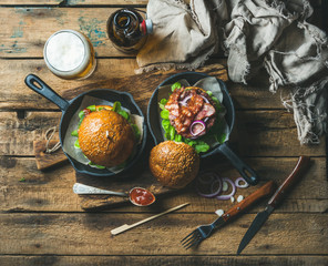 Wall Mural - Homemade beef burgers with crispy bacon and fresh vegetables in small cast iron pans and glass of wheat beer on rustic board over old wooden background, top view, horizontal composition