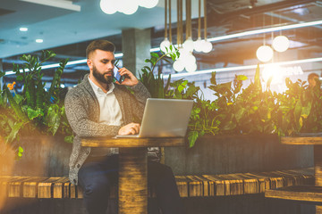 Wall Mural - Young
 bearded businessman, dressed in white shirt and gray cardigan, sitting 
at round wooden table in cafe and talking on cell phone while using 
laptop. House plants and round lamps.