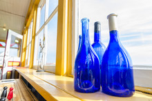 Blue Bottles In A Window Beside The Sea, Newfoundland Canada.  Wide Angle View, Blue Glass Bottles Decorate A Shelf At A Window With A View To The Atlantic In Grand Bank Newfoundland.