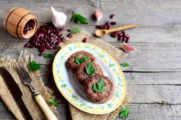 Wall Mural - Cutlets cooked from mashed red beans. Vegetarian cutlets, scattered raw red beans, garlic, fresh parsley, fork, knife, small wooden spoon and decorative barrel on old wooden background. Vintage style