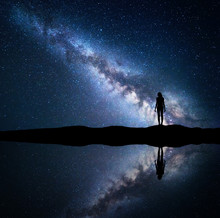 Milky Way. Night Starry Sky And Silhouette Of A Standing Woman On The Mountain Near The Lake With Sky Reflection In Water. Landscape With Blue Milky Way And Woman. Galaxy, Universe. Space Background