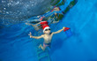 © alexbard - Happy child in a hat Santa Claus with a gift in hand swims underwater in the pool with outstretched arms on a blue background. The view from under the water. Landscape orientation