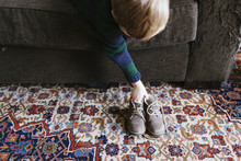 The Hands Of A Boy On A Dark Brown Couch And A Pair Of Little Boys Shoes On A Colourful Persian Rug.