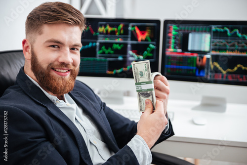Fotografía Cheerful bearded young businessman sitting and holding dollars