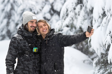 Young Man Couple Taking Selfie Photo In Snow Forest Outdoor Guys Holding Hands Winter Pine Woods