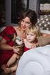 © romanovad - Women with girl sitting on a chair. Portrait of mother and daughter in identical red dresses near Christmas tree with gold decor