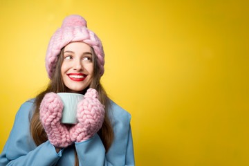 Woman in colorful winter clothes holding a cup of hot drink standing on the yellow background. Happy winter concept