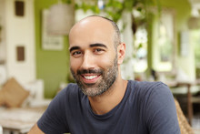 Close Up Shot Of Handsome Caucasian Man With Beard Dressed In T-shirt Looking And Smiling At Camera With Happy Cheerful Expression, Sitting At Sidewalk Restaurant On Sunny Day, Waiting For Friends