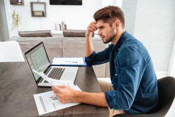 Concentrated man using laptop and looking on papers