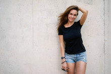 A Brunette Female With Long Hair Wearing A Blank Black T-shirt Is Smiling At The Camera While Standing On A Concrete Wall Background On A Street. Empty Space For Text Or Design.