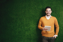 Attractive Man Standing Over A Green Grass Wall, Holding A Tablet 
