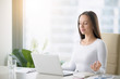 © fizkes - Young woman near the laptop, practicing meditation at the office desk, in front of laptop, online yoga classes, taking a break time for a minute, healing from paperwork and laptop radiation