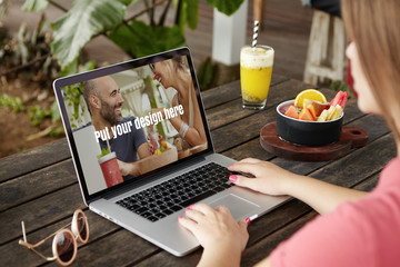 Rear shot of student girl in front of open laptop computer with hands on keyboard, learning online and browsing internet during breakfast, sitting at wooden table with shake, fruits and sunglasses