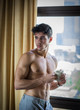 © theartofphoto - Sexy handsome young man standing shirtless in his bedroom next to window curtains, holding a coffee cup