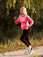 Happy pregnant woman jogging outside. Smiling expectant blonde running in forest. Sport, pregnancy, healthy lifestyle concept