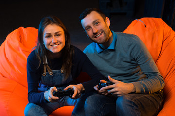 Two young gamer sitting on poufs and playing video games togethe