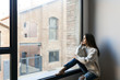 © Westend61 - Young woman sitting at a window looking out
