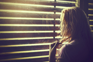 Sad and lonely blonde woman with wet hair looking through window blinds into the sunlight