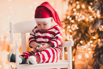 Cute baby girl under 1 year old sitting in chair holding Christmas ball in room over Christmas lights. Holiday time.