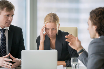 Young stressed businesswoman suffering from headache at important business meeting. Looking with doubtful expression at laptop screen, disagree with proposal or contract conditions, colleagues arguing