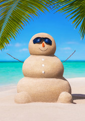 Wall Mural - Sandy snowman in black sunglasses and carrot nose at tropical ocean beach under palm leaves. Happy New Year and Merry Christmas travel destinations concept