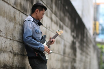 A young man wearing jeans, playing a guitar on a brick wall back