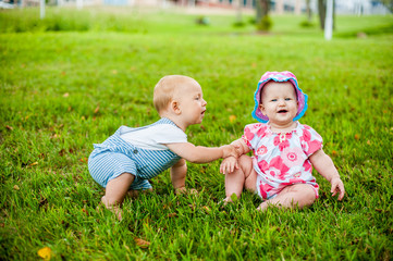 Two happy baby boy and a girl age 9 months old, sitting on the grass and interact, talk, look at each other.