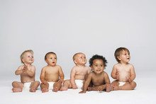 Row Of Multiethnic Babies Sitting Side By Side Looking Away Isolated On Gray Background