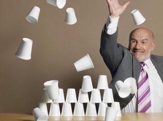 Middle aged businessman knocking down pyramid of plastic cups against brown background