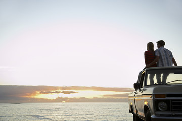 Rear view of young couple on pick-up truck parked in front of ocean enjoying sunset