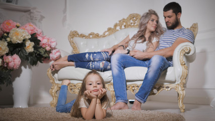 Close-up of happy family, expecting new baby, with little daughter in the foreground, lying on the carpet, dangling her legs and smiling. Her father and pregnant mother