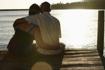 Wall Mural - Rear view of happy senior couple sitting on edge of pier by lake