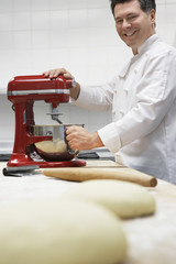 Wall Mural - Side view of a smiling male chef using dough mixer in kitchen with focus on dough in foreground