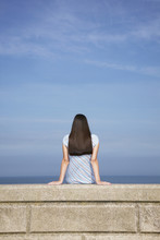 Rear View Of Young Woman Sitting On Stone Ledge At Beach Against Blue Sky