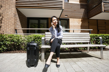 Wall Mural - Full length of businesswoman answering cell phone while sitting by luggage on bench against building