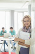 © moodboard - Portrait of beautiful businesswoman holding files with colleagues working in background at creative office