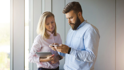 Bearded businessman in blue shirt and business woman with blonde hair in pink shirt standing near window. Man showing woman information on smartphone screen. Girl holding tablet computer.