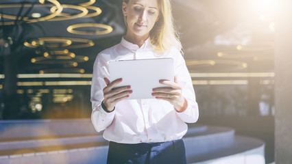 Wall Mural - Front view. Young smiling businesswoman with blonde hair in pink shirt standing in room with modern interior and uses tablet computer. Girl holding digital tablet and looking at its screen.