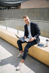 Wall Mural - Young hipster businessman in suit typing laptop near office outd