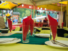 Picture Of The Dining-table Served With Red Napkin. White Plates With Flatware And Red Napkin Against The Blurred Background Of The Guests Of A Restaurant. Plates, Flatware, Glass And Red Napkin.