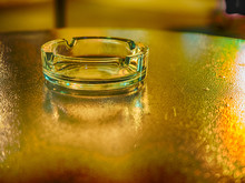 Picture Of The Glassy Ashtray Close Up. Ashtray Laying On The Blurred Surface Of The Table. Blurred Background Of The Ashtray On The Table. Blurred Surface Of The Table Reflects Yellow Sunlight.